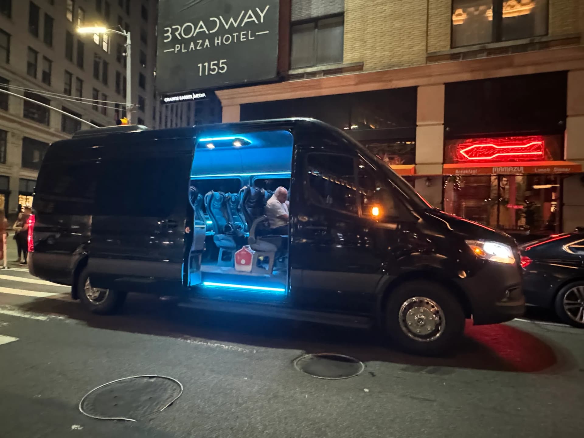 Black Sprinter limo van with blue interior lighting waiting outside a Broadway hotel at night.