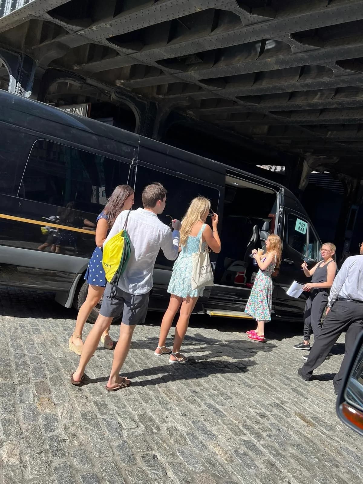 Group of passengers boarding a black Mercedes Sprinter van on a cobblestone NYC street beneath an elevated railway.
