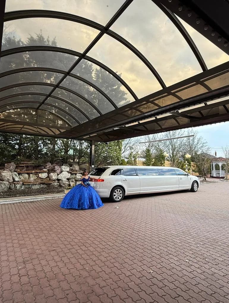 Quinceanera in a blue gown beside a white stretch limousine at the venue entrance