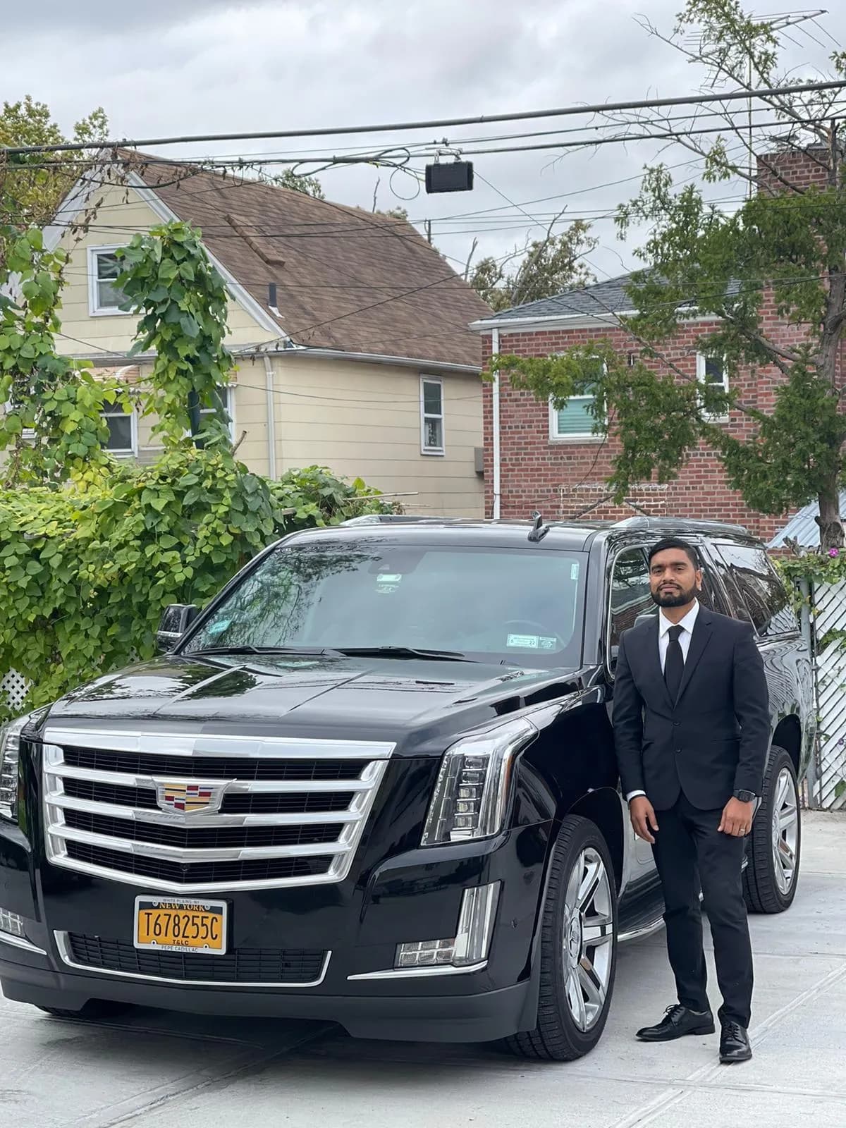 Chauffeur in a black suit beside a Cadillac Escalade in a Queens driveway