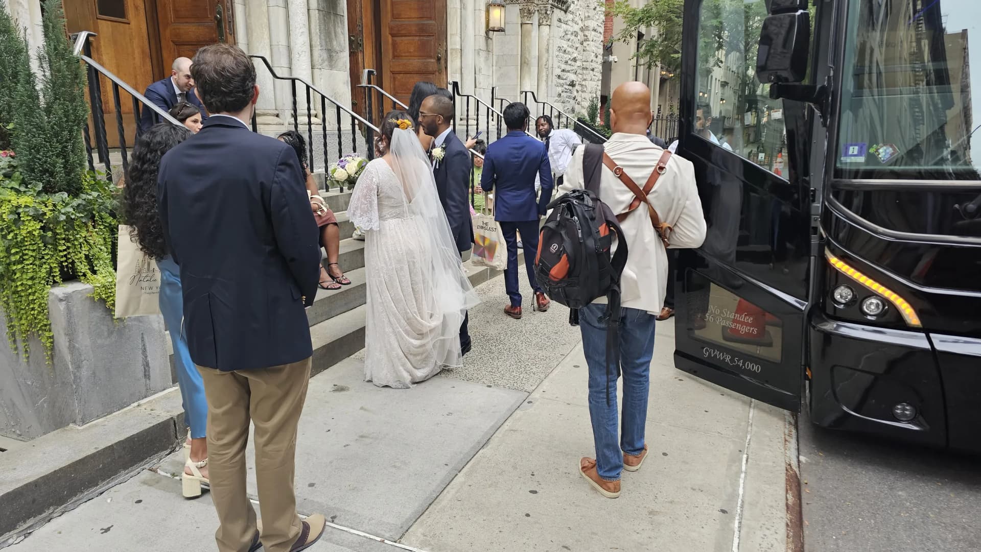 Bride and groom kiss on the church steps as guests head to the coach bus