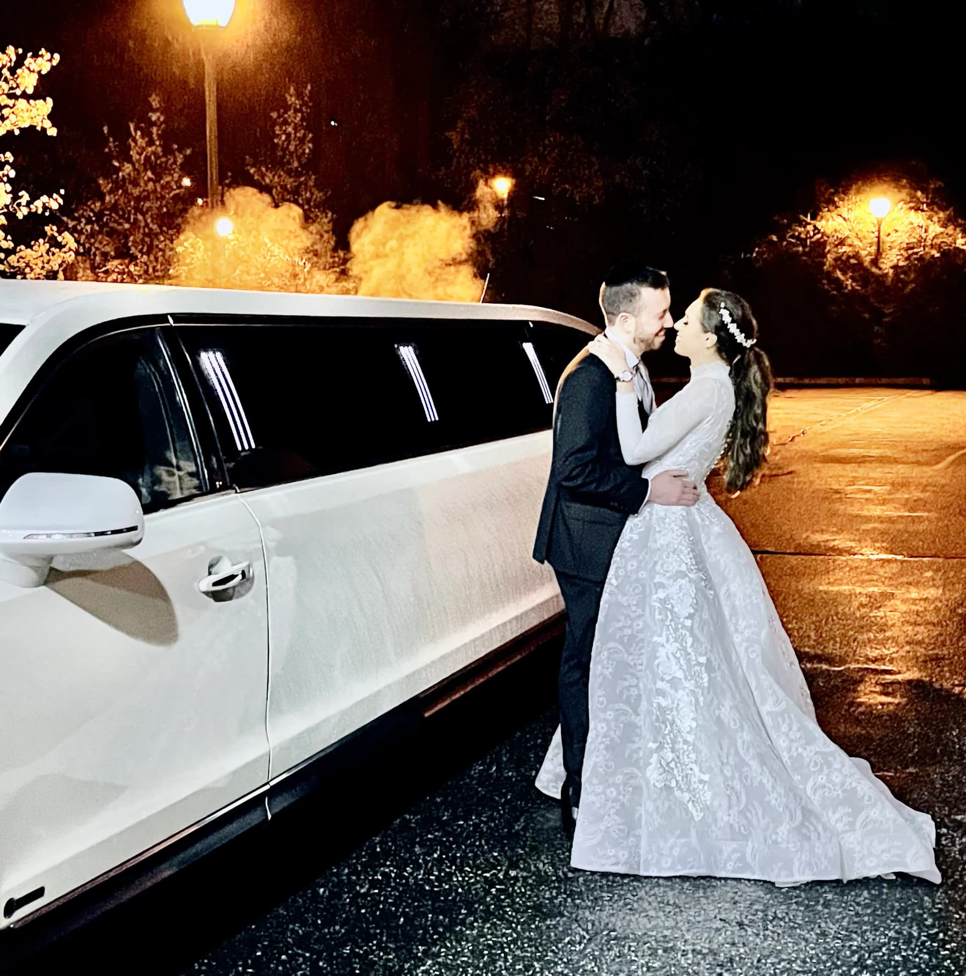 Newlyweds share a moment by their white stretch limousine after dark